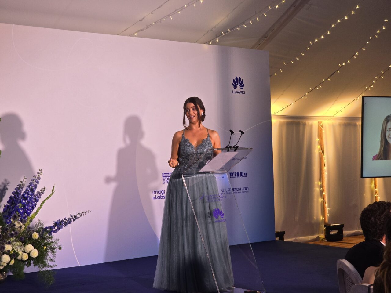 Huawei Summer School for Female Leadership Farewell Gala. Kobieta w eleganckiej sukni przemawia na scenie podczas gali, obok jest ekran z wyświetlonym wizerunkiem innej kobiety.