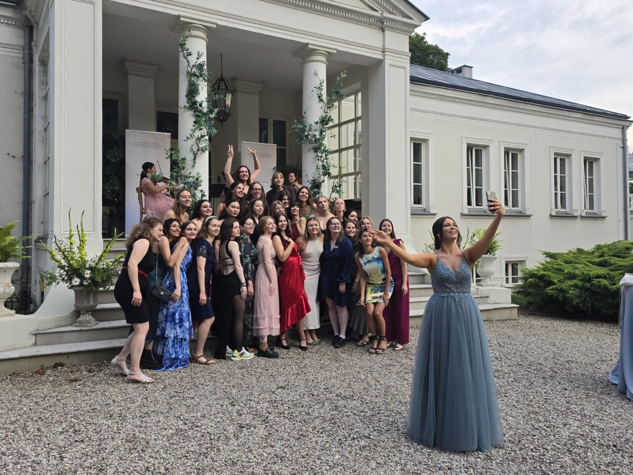 Grupa młodych kobiet na zewnątrz klasycznego białego budynku, jedna z nich robi selfie.