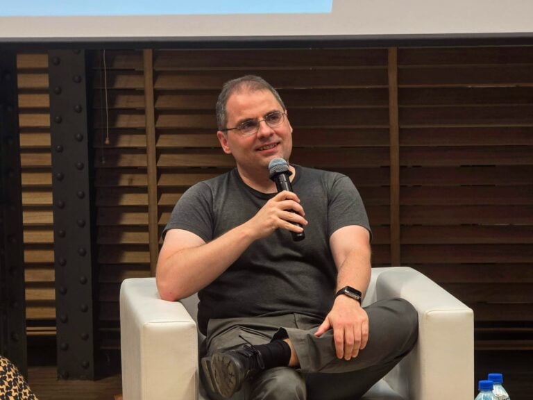 Debata IDEAS NCBR z udziałem Aleksander Mądry z OpenAI. Mężczyzna siedzący na białym fotelu, trzymający mikrofon, uśmiechający się, ubrany w szarą koszulkę i okulary.