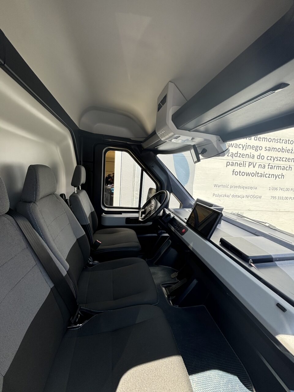 Wnętrze kabiny samochodu dostawczego z trzema miejscami siedzącymi, kierownicą oraz ekranem dotykowym na desce rozdzielczej.
