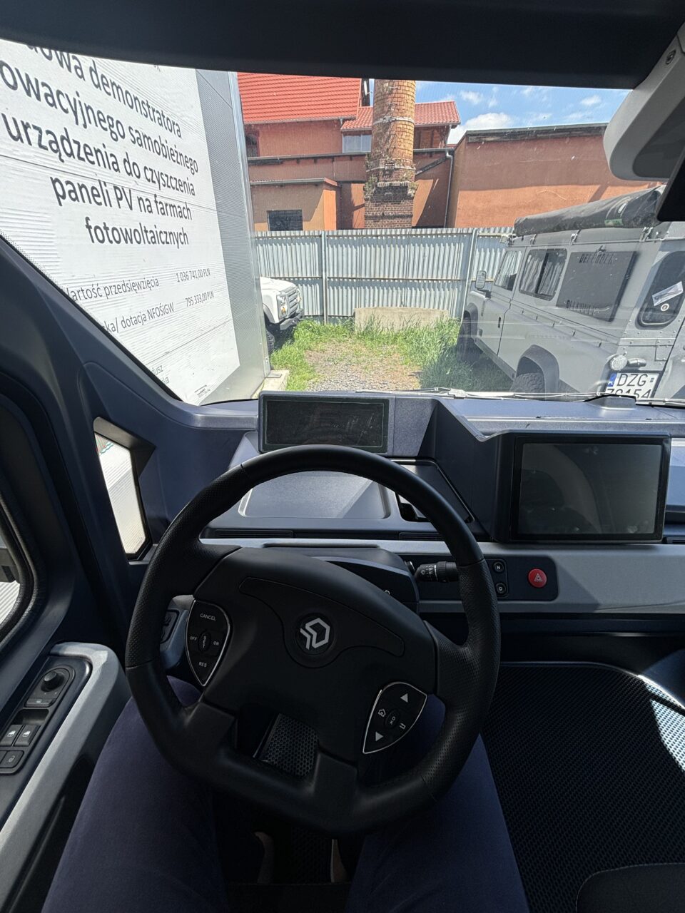 Widok wnętrza samochodu elektrycznego z kokpitu kierowcy z widoczną kierownicą i ekranami, przez przednią szybę widać zaparkowane samochody i budynek.