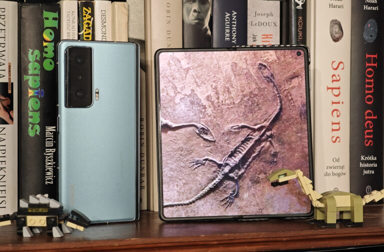 Smartfon HONOR na półce z książkami, obok otwartego składanego telefonu wyświetlającego obraz skamieniałości dinozaura oraz dwóch figurek LEGO dinozaurów.