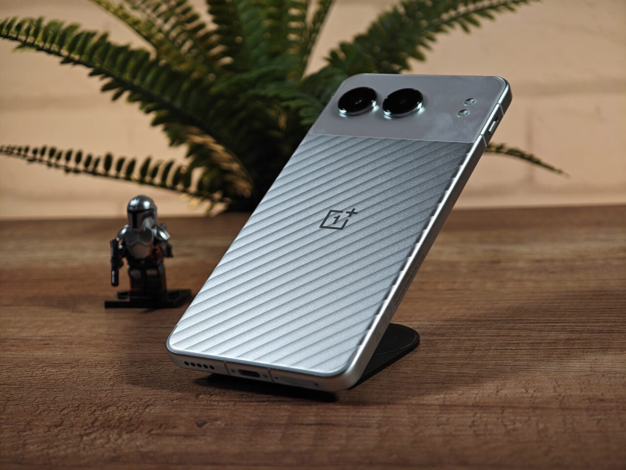 Srebrny smartfon OnePlus Nord 4 stojący na drewnianym biurku obok figurki Mandaloriana, w tle zielona paproć.