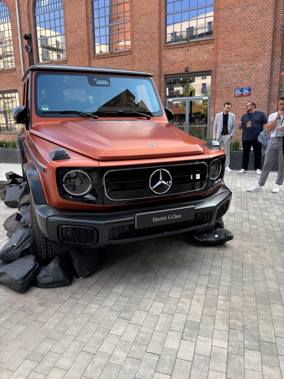 Samochód elektryczny Mercedes-Benz G-Class na wystawie przed budynkiem z cegły, w tle kilku mężczyzn.