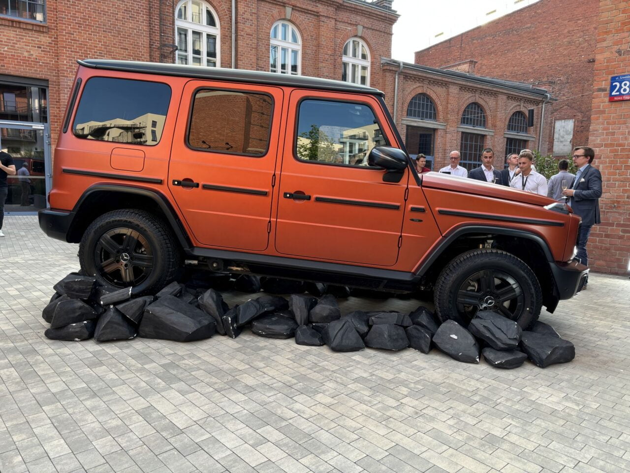 Pomarańczowy SUV zaparkowany na czarnych kamieniach na tle ceglanego budynku, ludzie stojący obok.