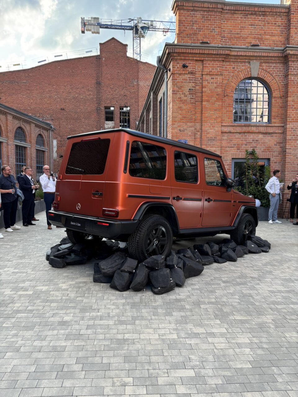 Brązowy samochód terenowy Mercedesa zaparkowany na czarnych skałach przed budynkiem z czerwonej cegły, ludzie stojący w tle.