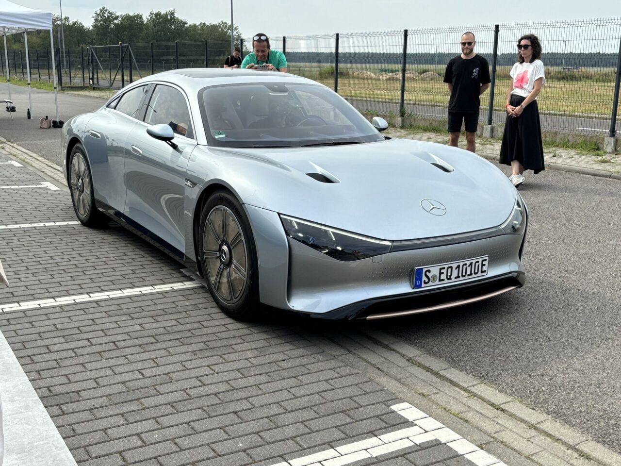 Srebrny samochód marki Mercedes-Benz EQX na parkingu, z dwoma osobami stojącymi w tle.