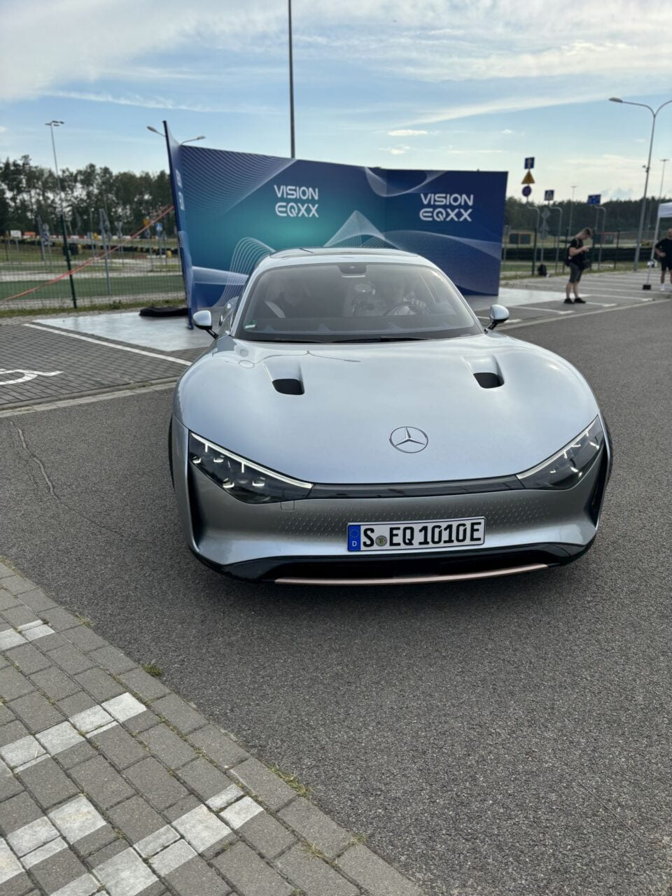 Srebrny samochód elektryczny Mercedes-Benz Vision EQXX zaparkowany na parkingu z banerem reklamowym w tle.