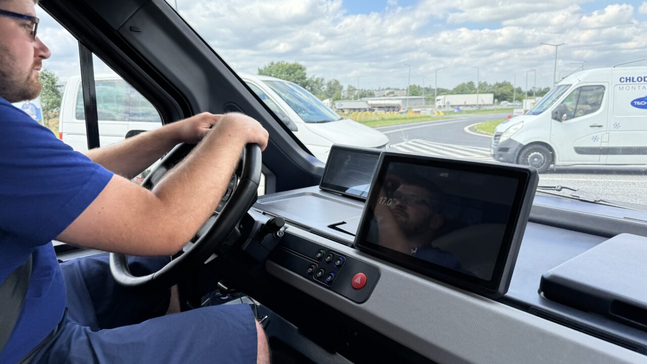 Kierowca w niebieskiej koszulce prowadzi pojazd, patrząc na drogę; we wnętrzu samochodu widoczne dwa ekrany, na drodze obok inny pojazd.
