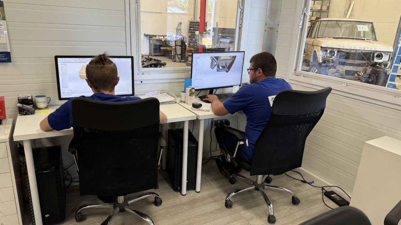 Dwóch mężczyzn w biurze pracujących przy komputerach; na ekranach wyświetlane są projekty 3D. W tle przez okno widać warsztat samochodowy i rozebrane auto.