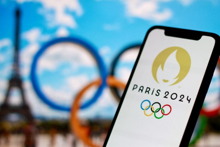 Logo Igrzysk Olimpijskich Paryż 2024 na ekranie telefonu na tle wieży Eiffla i pięciu pierścieni olimpijskich.