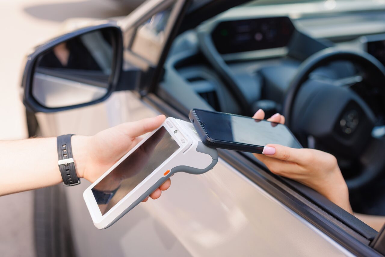 Kierowca płaci smartfonem z NFC, przykładając go do terminala płatniczego przez okno samochodu.