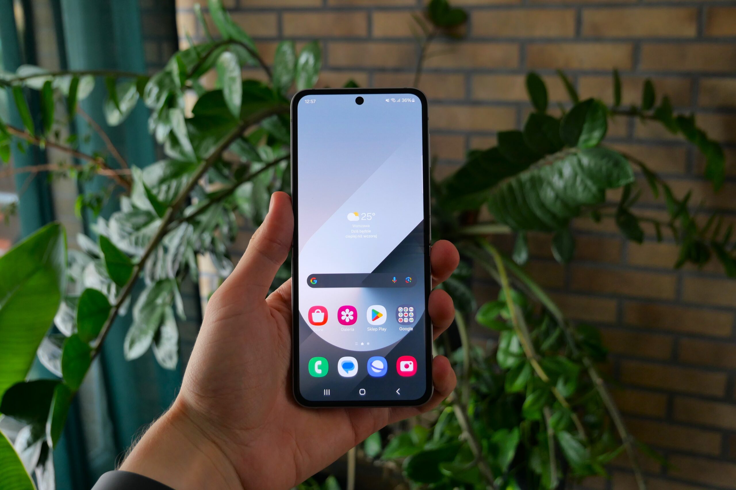Osoba trzymająca smartfon z ekranem głównym wypełnionym różnymi aplikacjami na tle zielonych roślin i ceglanej ściany. OneUI jest na tym urządzeniu zainstalowany.