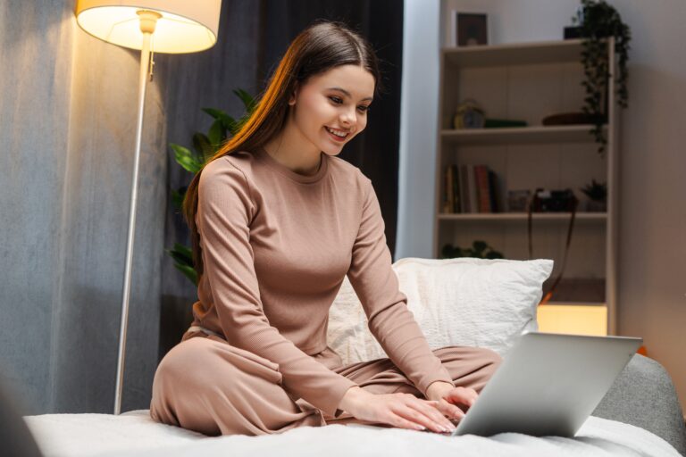 Młoda kobieta siedząca na łóżku z laptopem, ubrana w beżową piżamę, uśmiechnięta, w przytulnie oświetlonym pokoju.