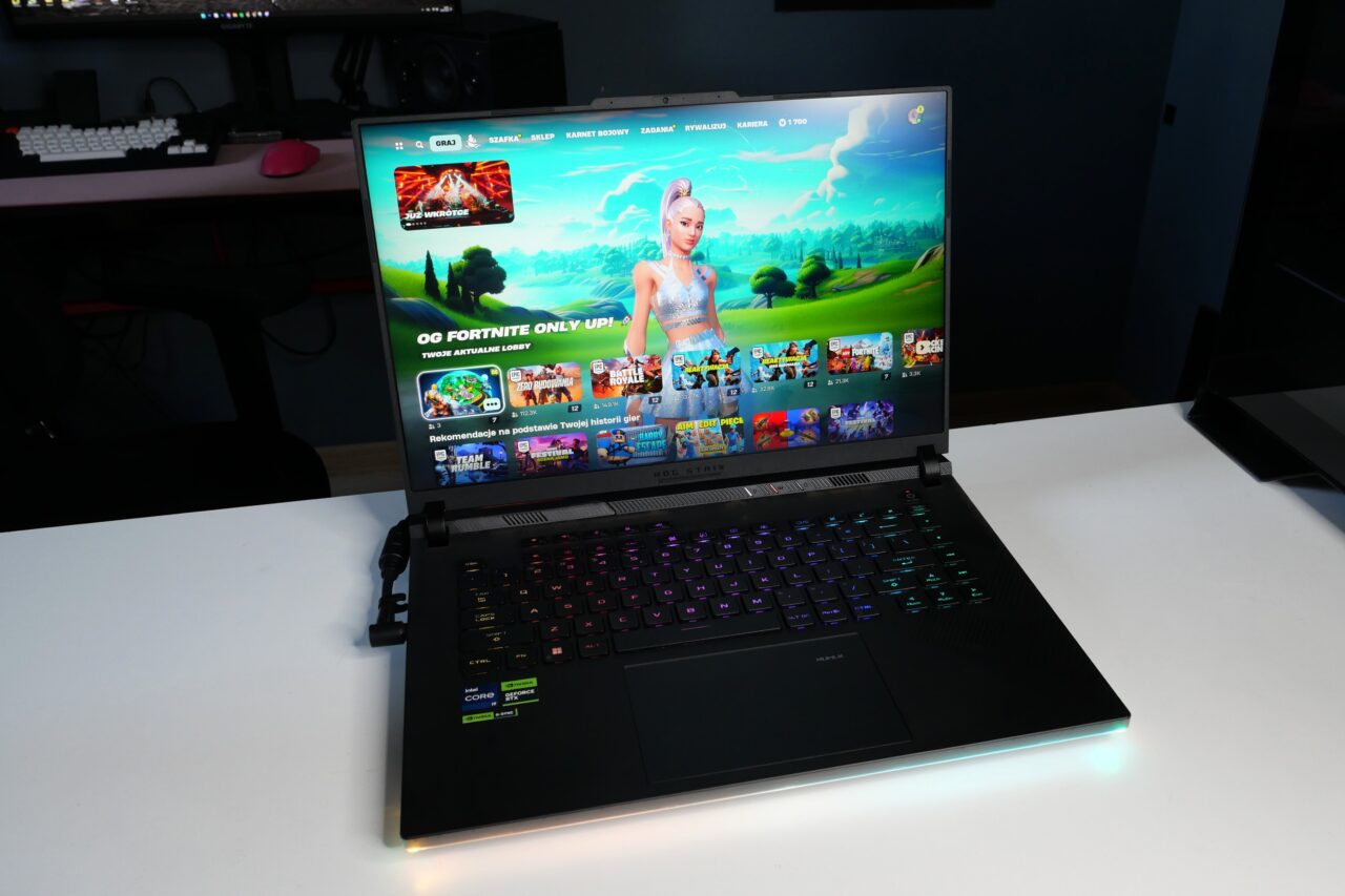 Laptop z uruchomioną grą Fortnite na ekranie, stoi na białym biurku. W tle widać klawiaturę i inne akcesoria komputerowe.