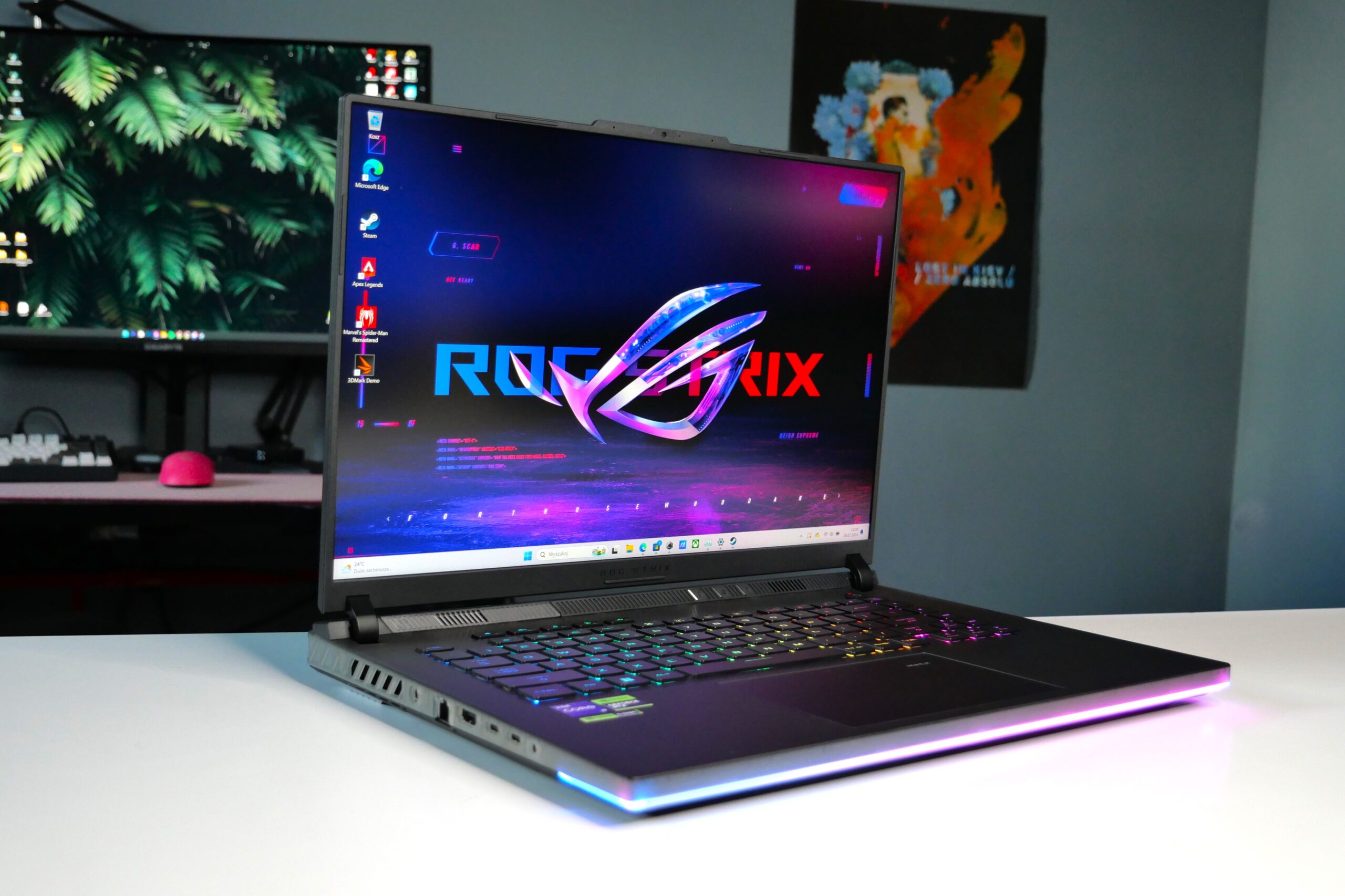 Laptop ASUS ROG Strix na biurku z włączonym ekranem i podświetlaną klawiaturą; w tle drugi monitor oraz klawiatura z myszką.