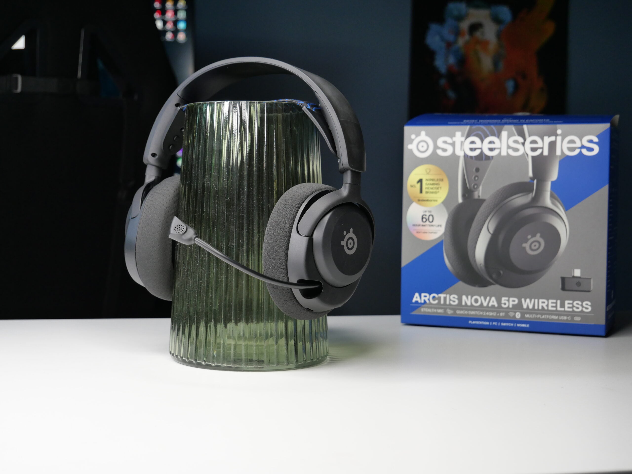 Słuchawki SteelSeries Arctis Nova 5P Wireless umieszczone na szklanym wazonie, w tle pudełko z ich opakowaniem.