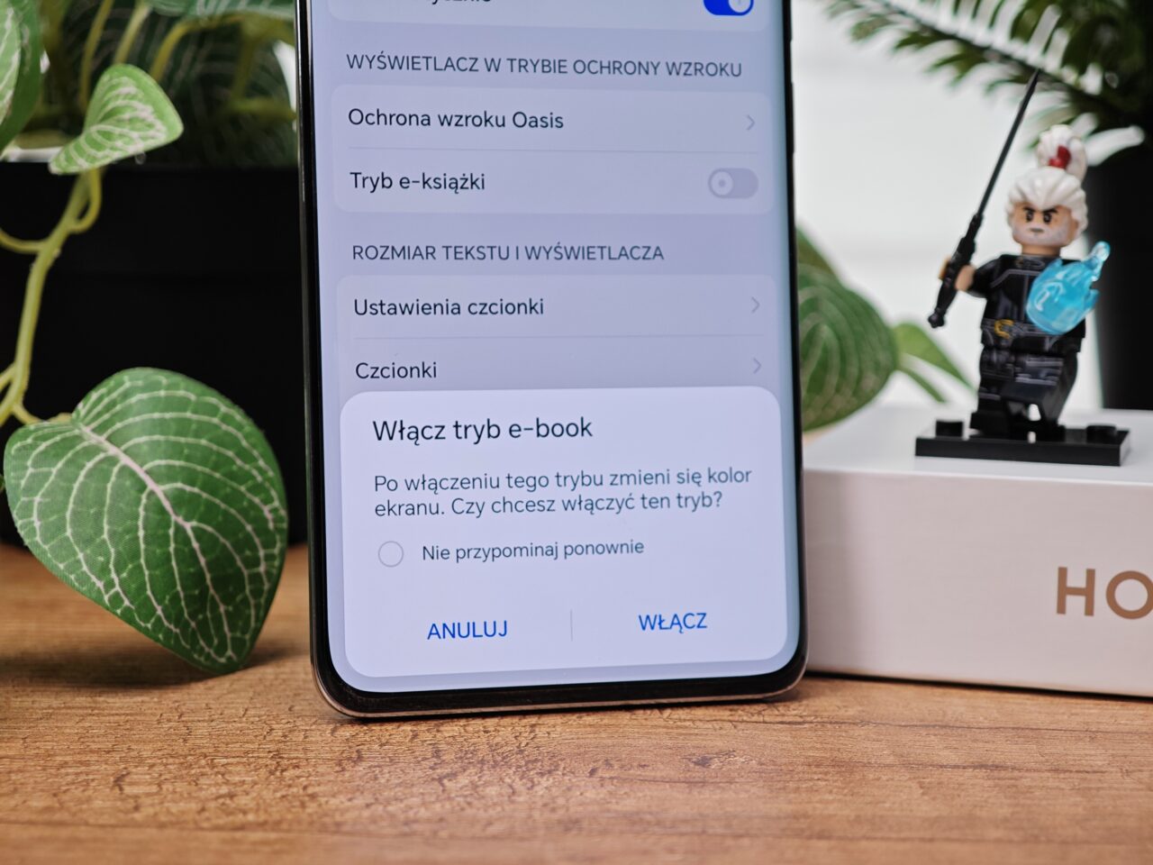 Ekran smartfona z ustawieniami, w tym opcją włączenia trybu e-book, roślina w tle oraz figurka ninja na pudełku.