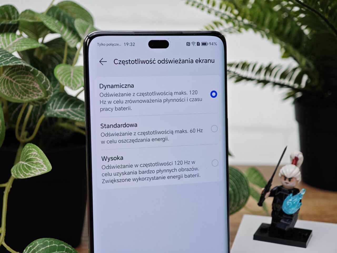 Ustawienia częstotliwości odświeżania ekranu na smartfonie z trzema opcjami: Dynamiczna, Standardowa, Wysoka; w tle rośliny i figurka postaci.