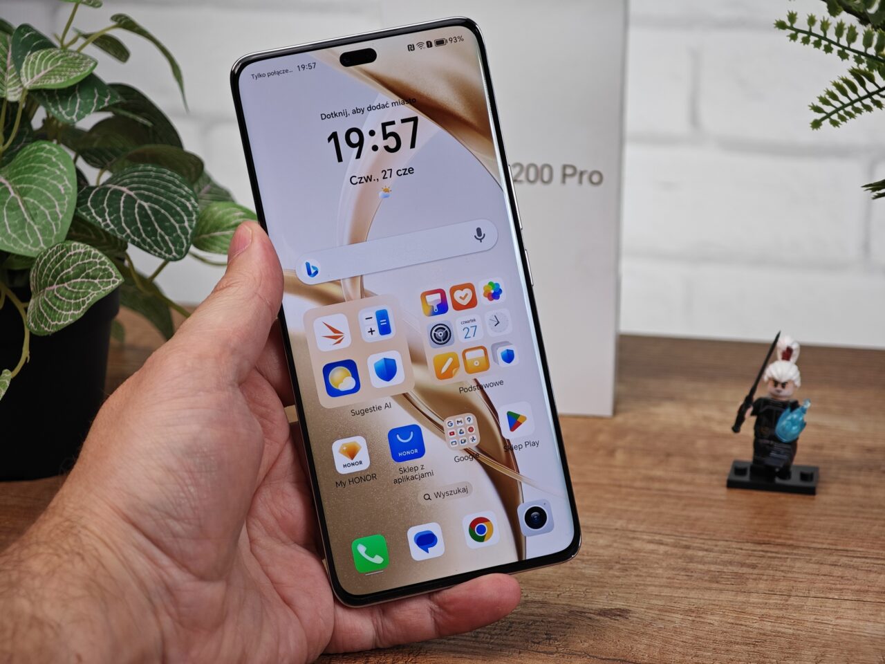 Osoba trzymająca w ręku smartfon HONOR 200 Pro z widocznym ekranem startowym, w tle roślina doniczkowa, figurka Lego i pudełko telefonu.