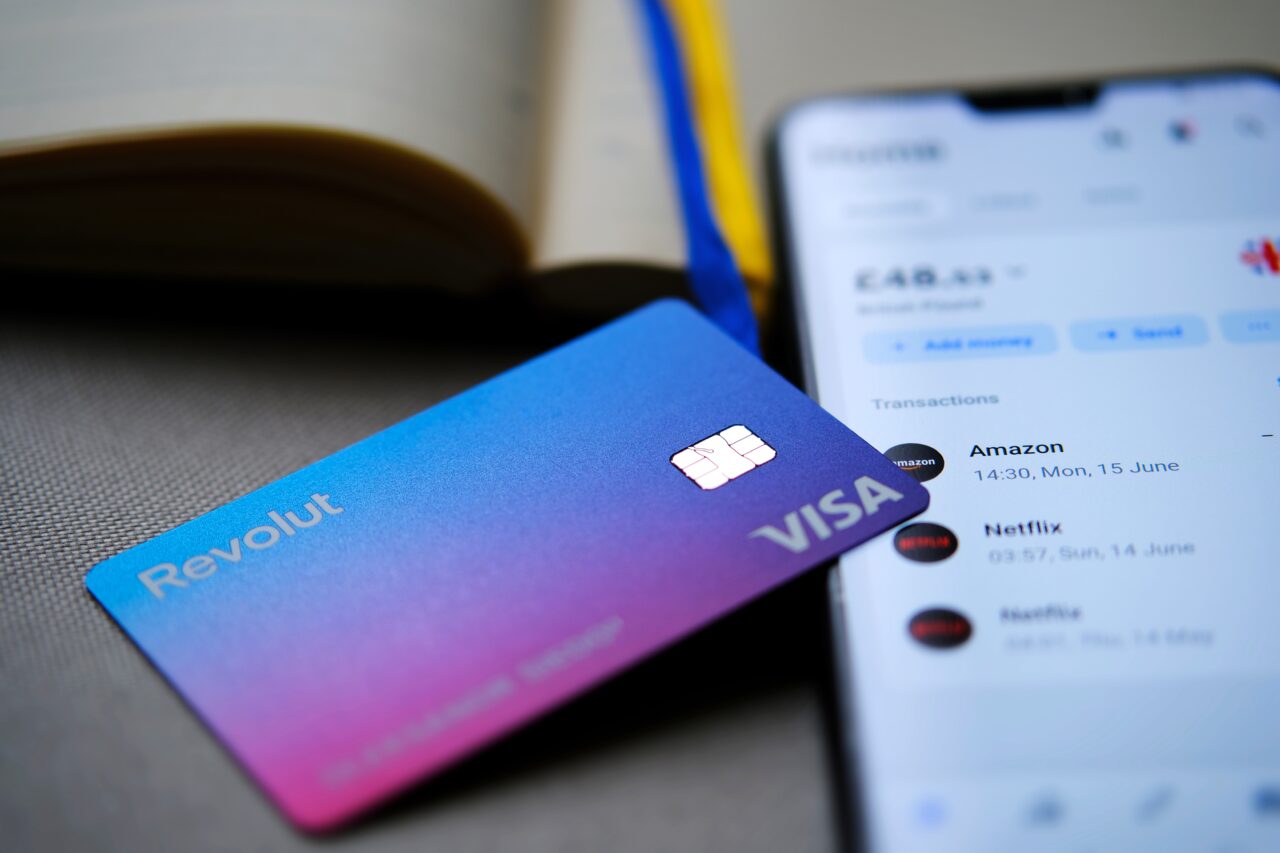 Karta Revolut VISA leżąca na stole obok otwartej książki i smartfona z wyświetloną aplikacją bankową.