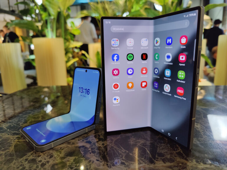 Składany smartfon Samsung Galaxy Z Fold6 i Flip6 na stole z ekranem głównym i dodatkowymi aplikacjami, w tle zielone rośliny i osoby.