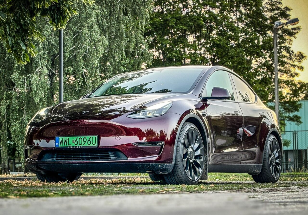 Bordowy samochód Tesla Model Y z polskimi, zielonymi tablicami rejestracyjnymi, zaparkowany na trawie, w tle drzewa.