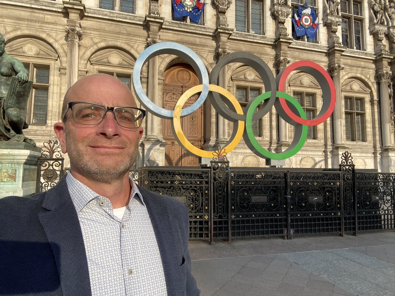 Mężczyzna w okularach robiący selfie przed budynkiem z symbolem olimpijskim.