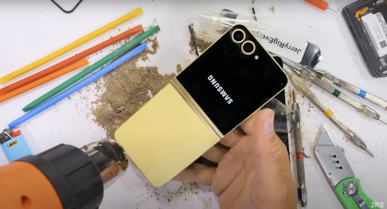 Smartfon Samsung Galaxy Z Flip na biurku otoczonym narzędziami, w tym wiertarka, zapalniczka Bic, kolorowe pałeczki i przyrządy do demontażu.