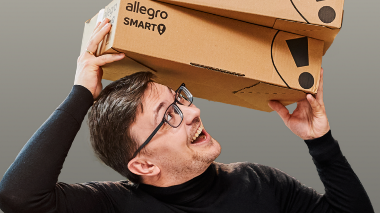 Mężczyzna trzymający dwa kartonowe pudełka Allegro Smart! na głowie, z uśmiechem na twarzy.