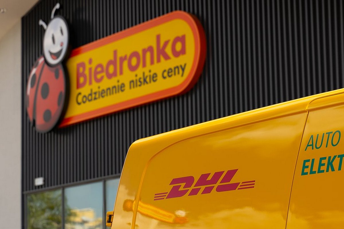 Logo sklepu Biedronka z napisem "Codziennie niskie ceny" oraz widoczny fragment żółtego samochodu DHL.