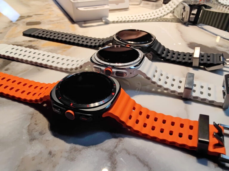 Trzy smartwatche z gumowymi paskami w kolorach: czarny, biały i pomarańczowy, leżące na marmurowym blacie.