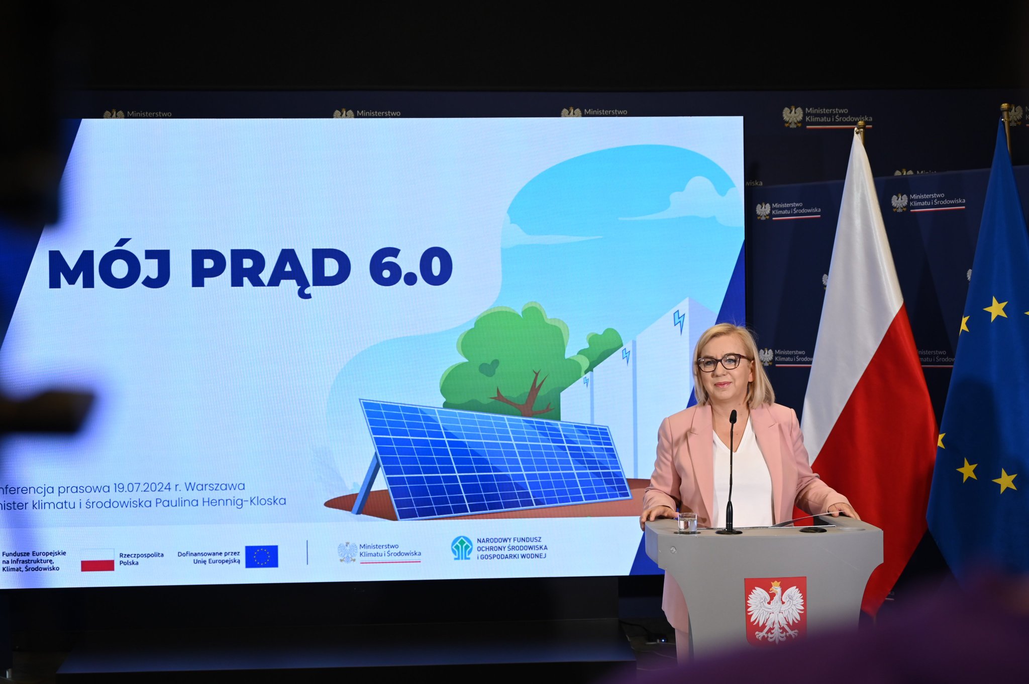 Minister klimatu i środowiska przy mównicy na konferencji prasowej "Mój Prąd 6.0" 19.07.2024 r. w Warszawie, na tle ekranu z panelem słonecznym i flagami Polski oraz Unii Europejskiej.