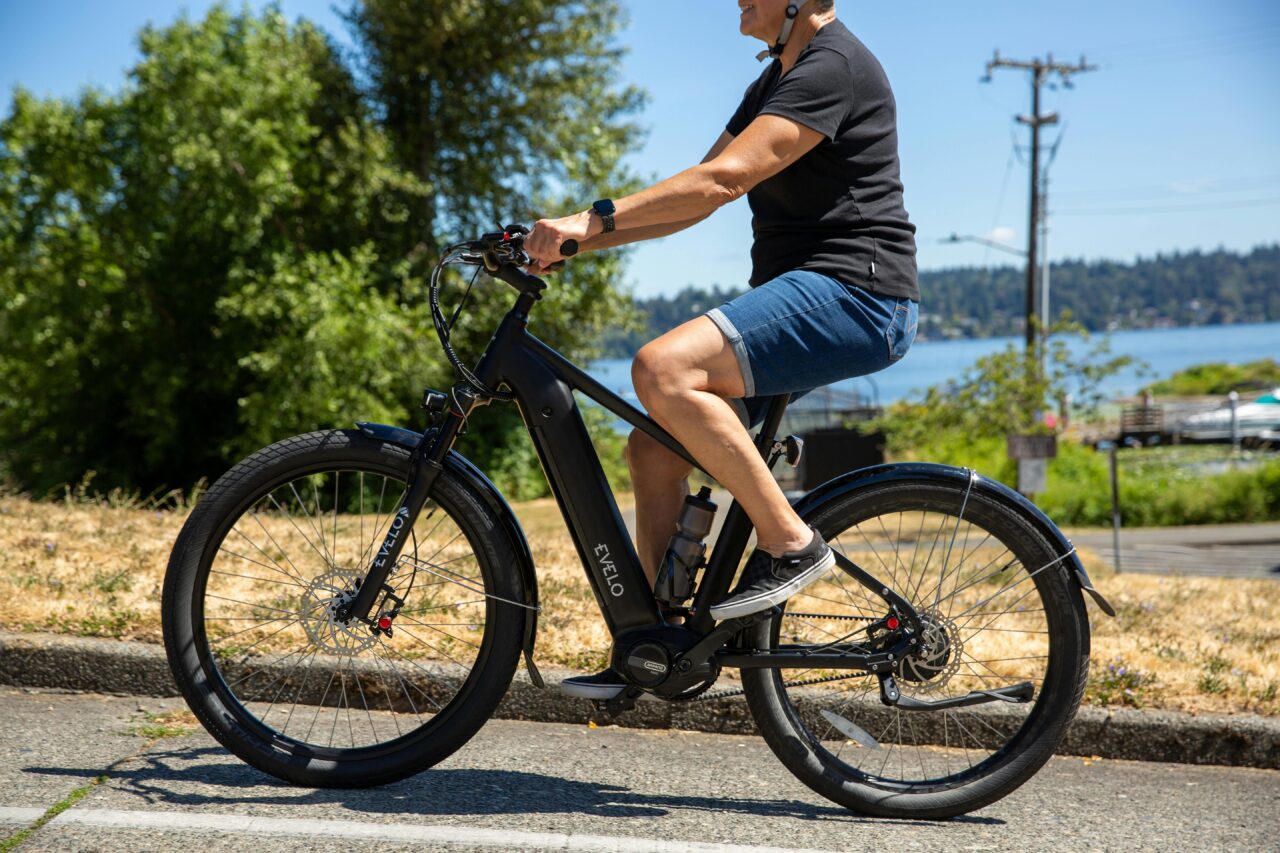 Mój rower elektryczny Osoba jadąca na czarnym rowerze elektrycznym Evelo po asfaltowej drodze, w tle drzewa i jezioro.