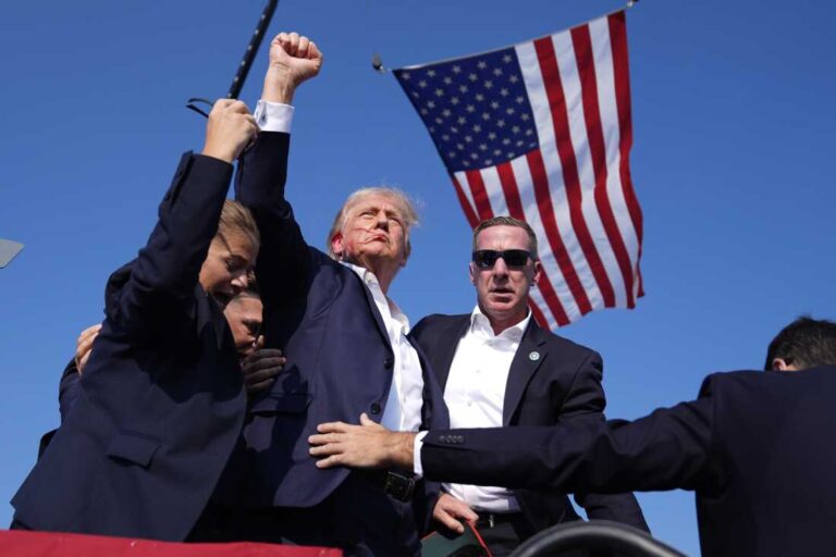 Grupa ludzi w garniturach pod amerykańską flagą, jedno z nich trzyma uniesioną pięść.