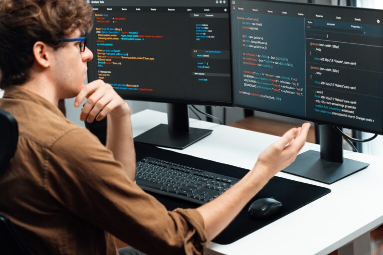 Mężczyzna pracujący przy komputerze z dwoma monitorami, na których widoczny jest kod programistyczny.
