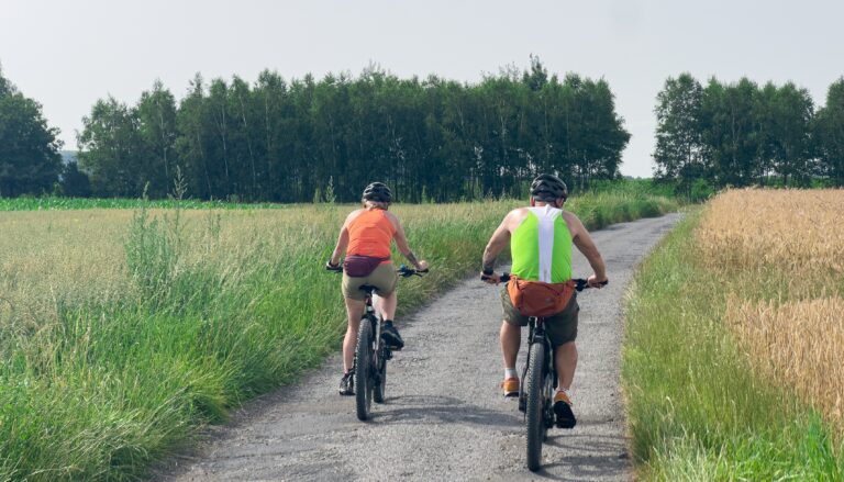 Dwóch rowerzystów jadących wąską ścieżką przez pola i las.