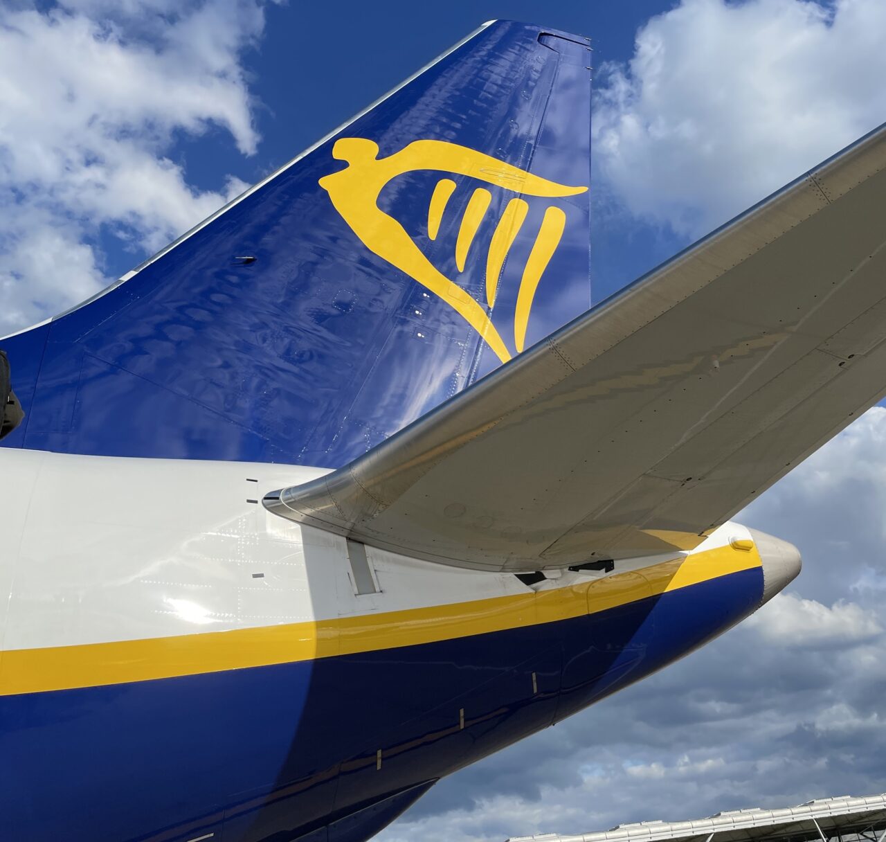 Ogon samolotu Ryanair z logo harfy, na tle nieba z chmurami.