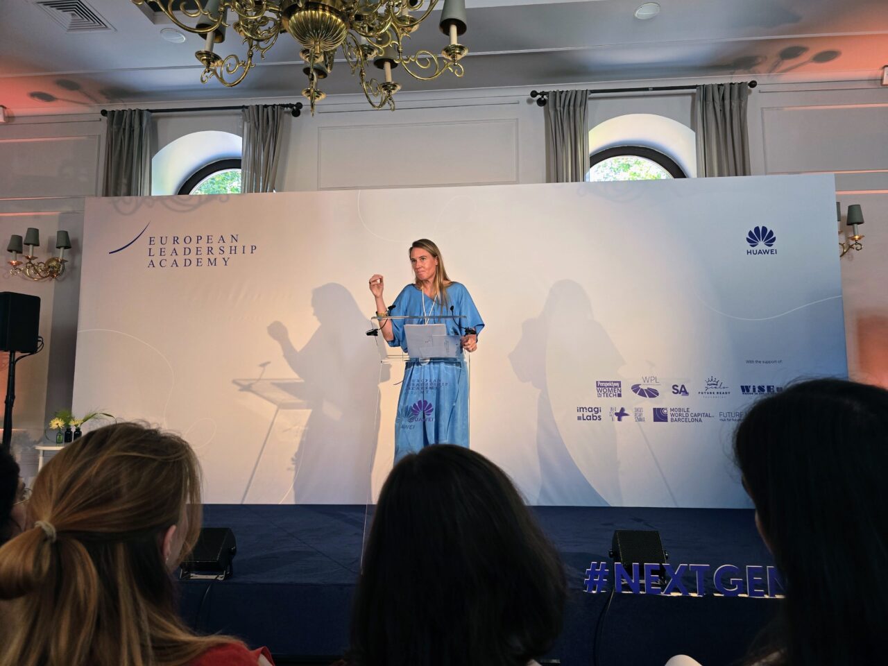 Huawei Summer School for Female Leadership in the Digital Age. Kobieta w niebieskiej sukni przemawia na scenie podczas wydarzenia European Leadership Academy, obok logo Huawei i hashtagu #NEXTGEN.