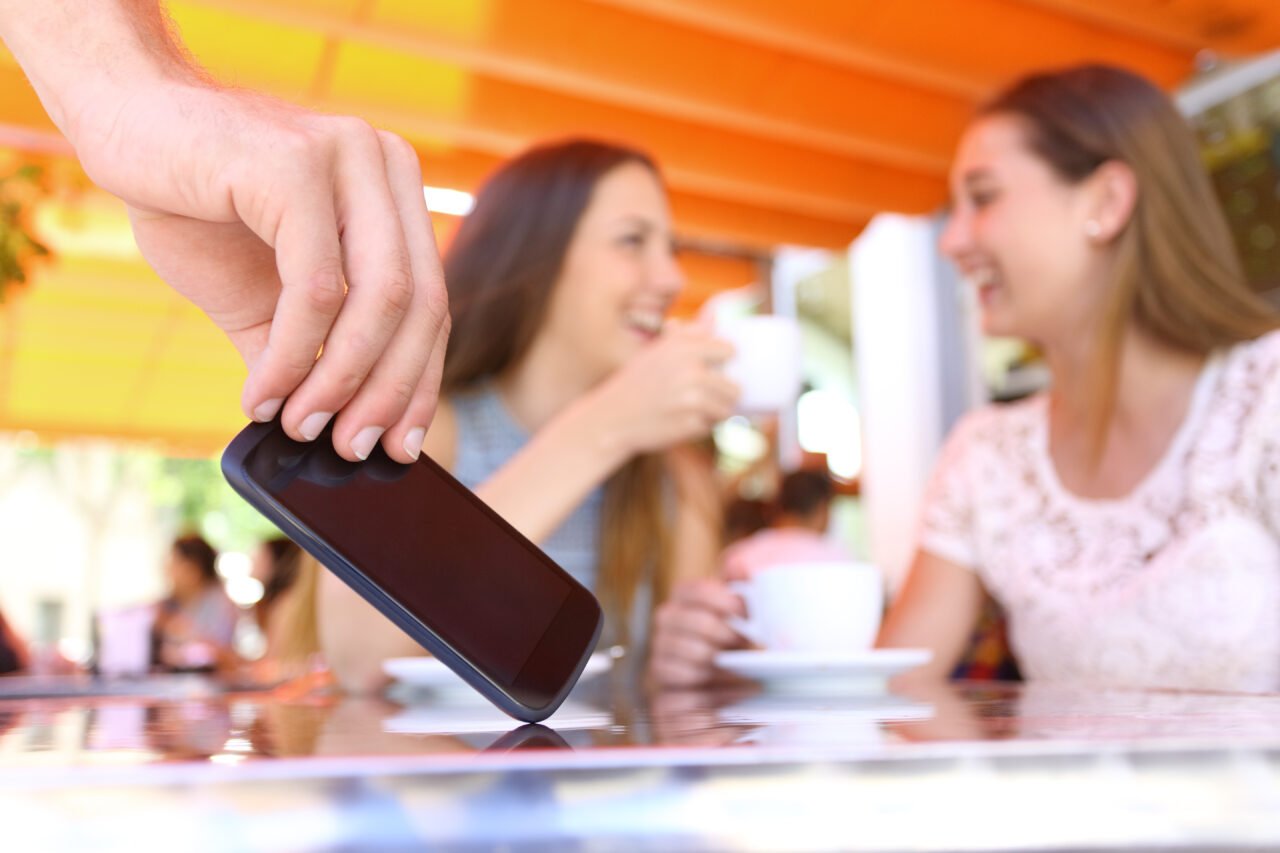 Ręka podnosząca telefon leżący na stole, w tle dwie kobiety pijące kawę i rozmawiające.