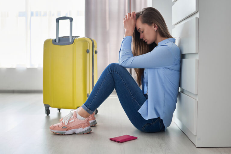 Kobieta siedzi na podłodze oparta o szafkę, z rękami na głowie, obok niej żółta walizka i różowy paszport.