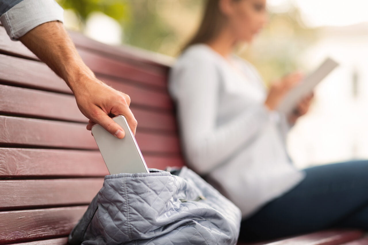 Kradzieże telefonów. Złodziej kradnie telefon z torby na ławce, kobieta w tle używa tabletu.