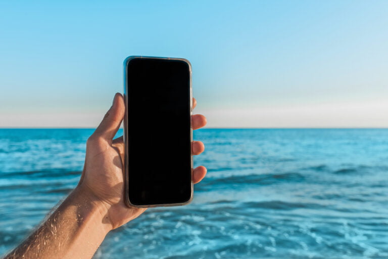 Ręka trzymająca smartfon na tle morza i bezchmurnego nieba. Temperatura wody jest nieznana, ale w jej ustaleniu może pomóc specjalna aplikacja.
