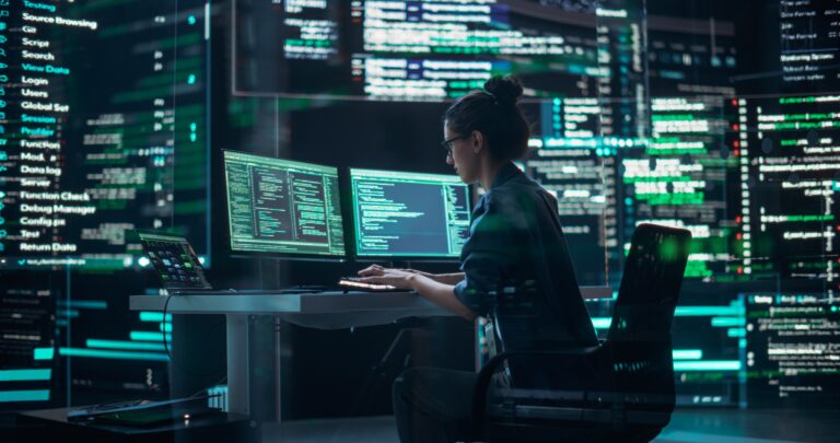 Kobieta siedząca przy biurku przed kilkoma monitorami, pracująca nad kodowaniem w ciemnym pokoju z wyświetlanymi na ścianach liniami kodu.