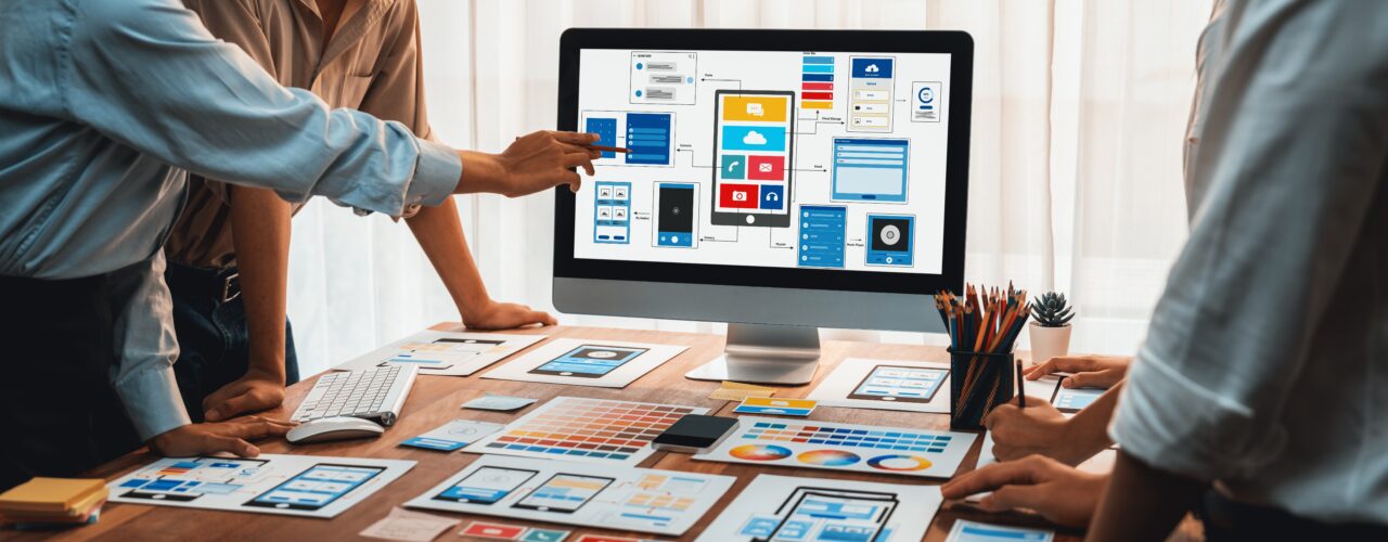 Praca w UX i UI. Zespół projektujący interfejs użytkownika aplikacji mobilnej na komputerze, wokół stołu z rozłożonymi planami i próbnikami kolorów.
