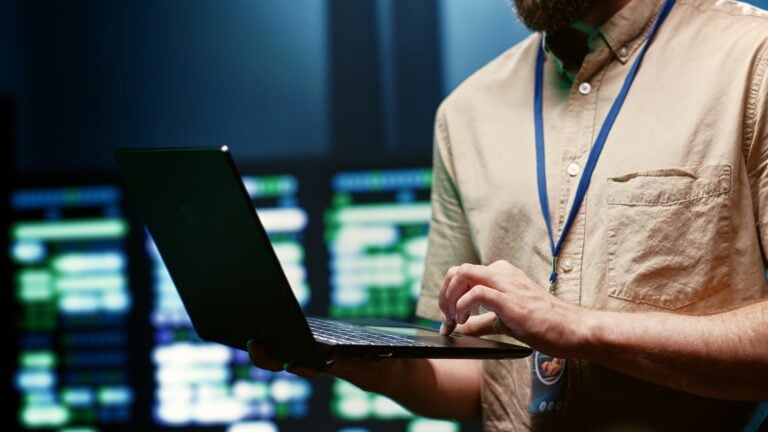 Odzyskiwanie danych. Osoba trzymająca laptop w rękach, w tle rozmyte ekrany komputerowe z zielonymi i niebieskimi wyświetleniami.