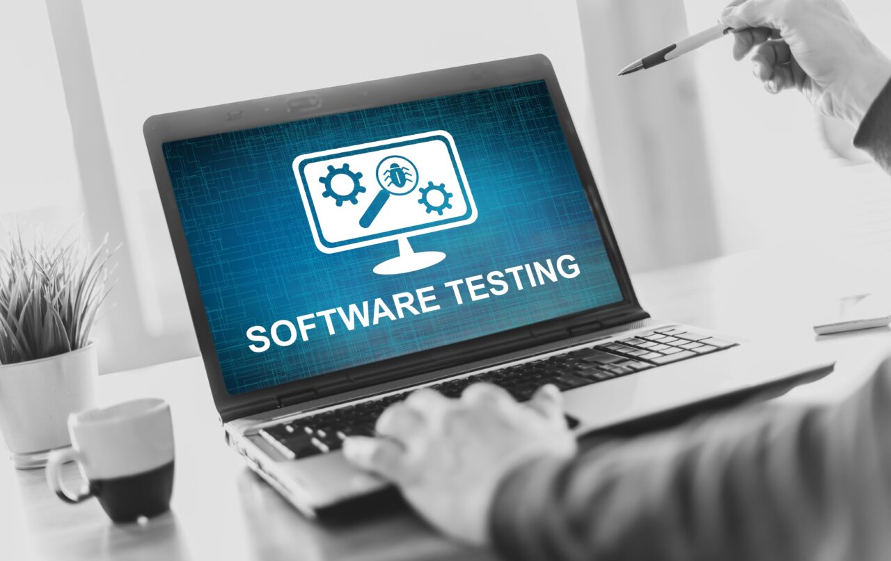 Tester oprogramowania. Laptop z wyświetlonym ekranem "SOFTWARE TESTING", na biurku kubek i doniczka z rośliną, osoba trzyma długopis.