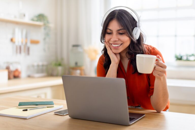 Praca zdalna. Kobieta w słuchawkach, trzymająca kubek z napojem, siedzi przy stole przed laptopem, uśmiechając się.