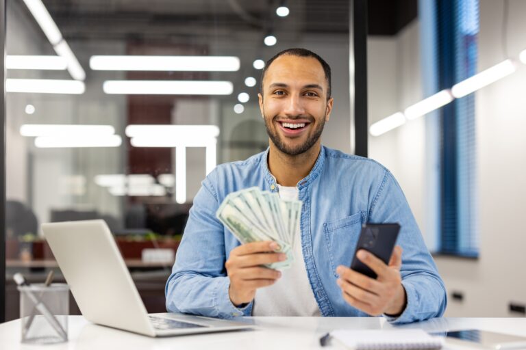 Mężczyzna z brodą w dżinsowej koszuli siedzi przy biurku w nowoczesnym biurze, trzymając w jednej ręce telefon, a w drugiej plik banknotów.