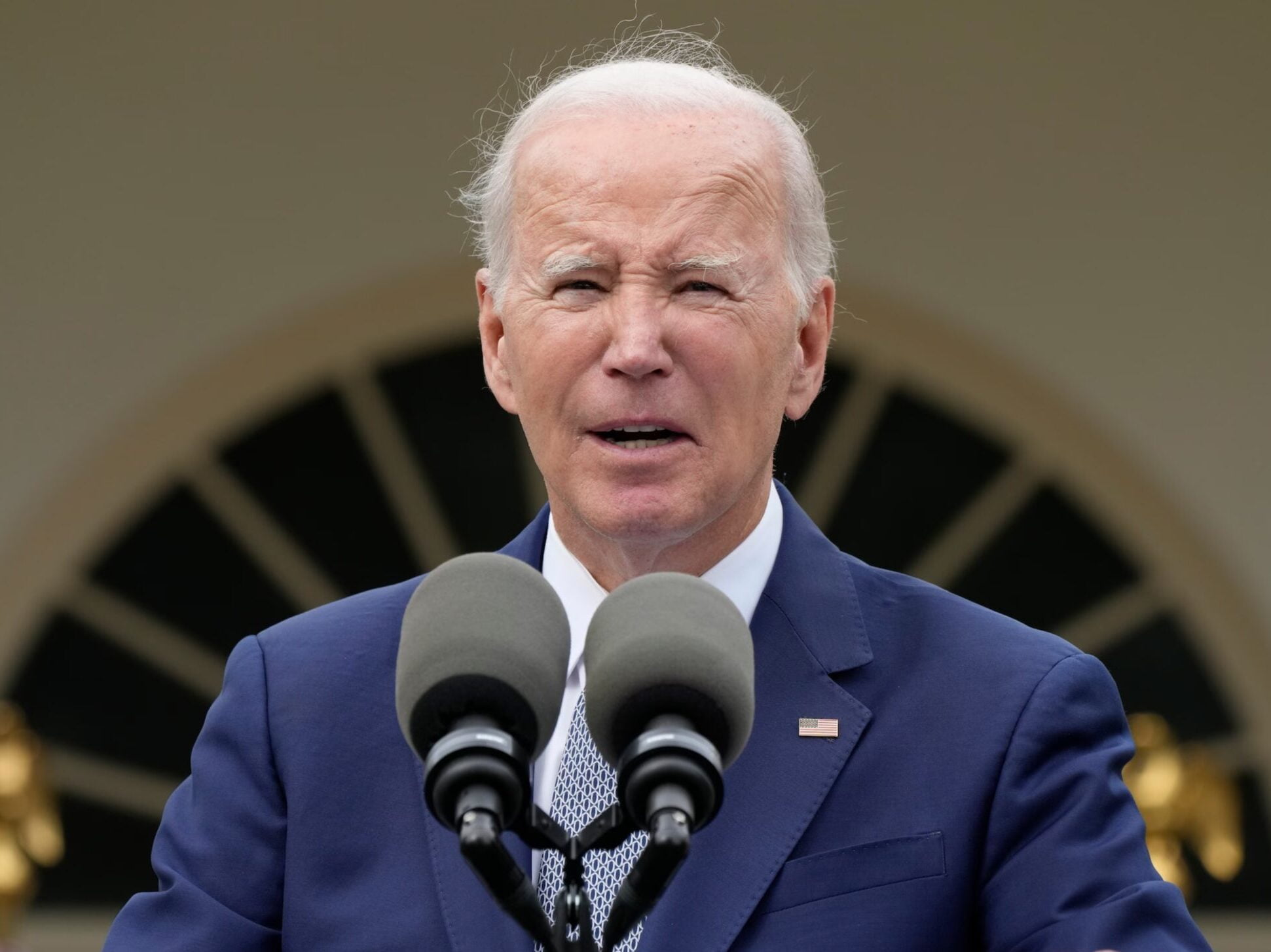 Joe Biden. Starszy mężczyzna w granatowym garniturze przemawia do mikrofonów na zewnątrz budynku.